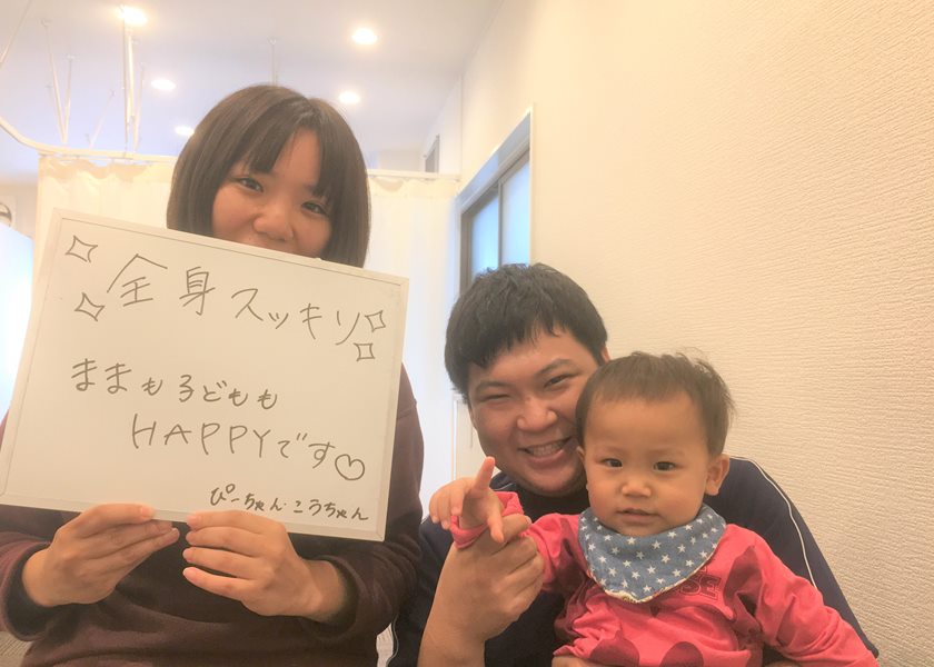 「全身スッキリ ままも子どももHAPPYです」と書かれたボードを持つ女性と、お子さんを抱いた院長のツーショット写真